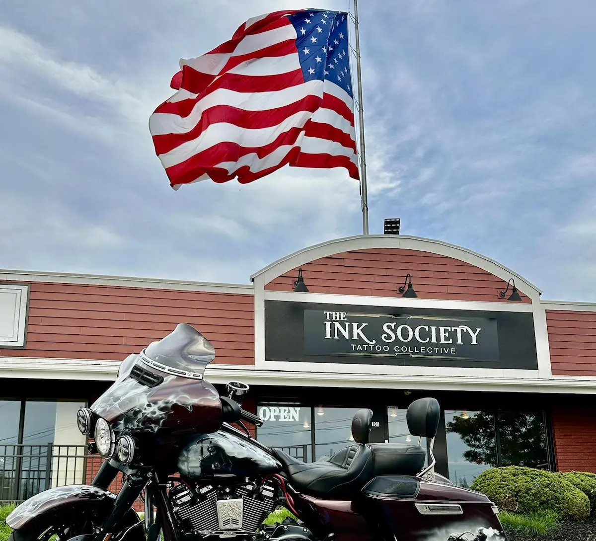 Harley-Davidson motorcycle parked in front of a tattoo and piercing studio with a large American flag displayed above the shop